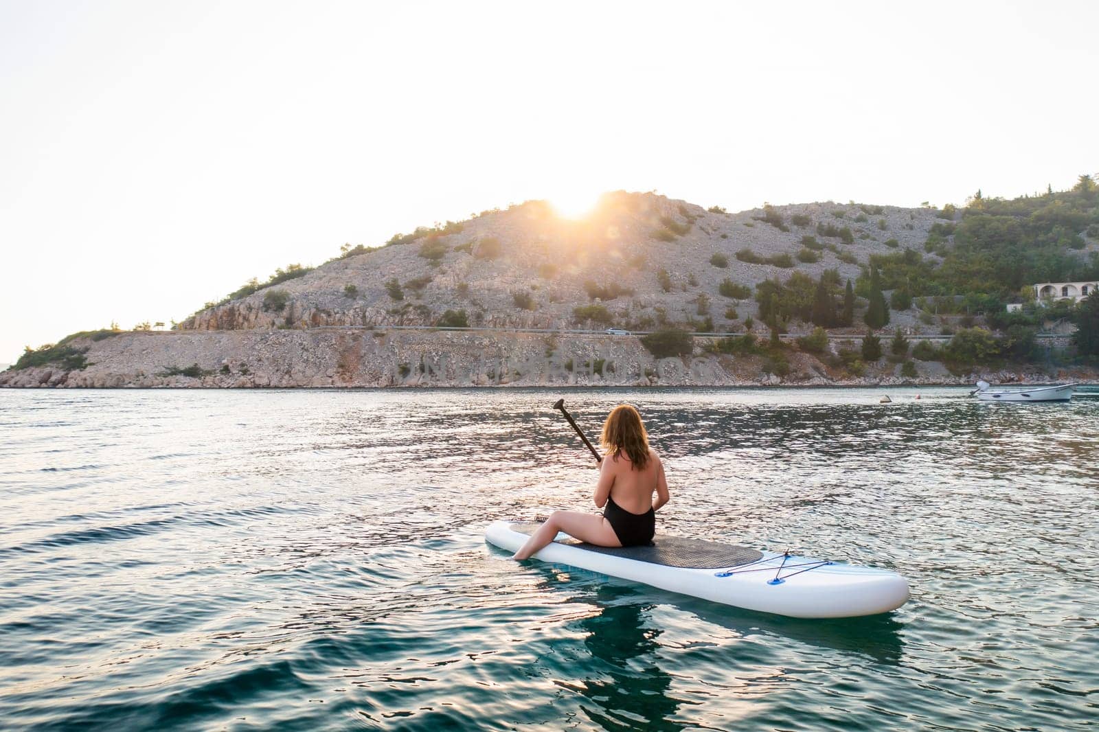 Woman paddle boarding at sunset on tranquil water near a mountain shoreline.