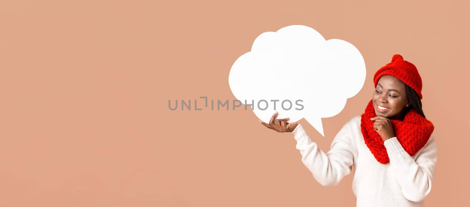 Pretty interested woman in winter hat holding communication bubble, touching her chin, beige studio background