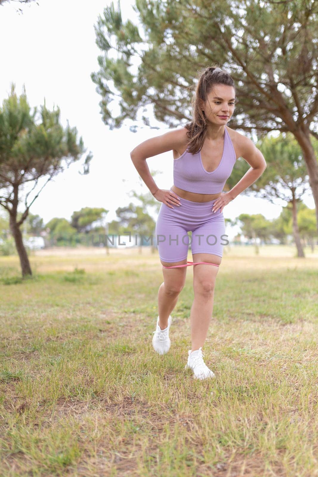 A young woman wearing a purple sports bra and shorts performs leg exercises with a resistance band in a grassy park.