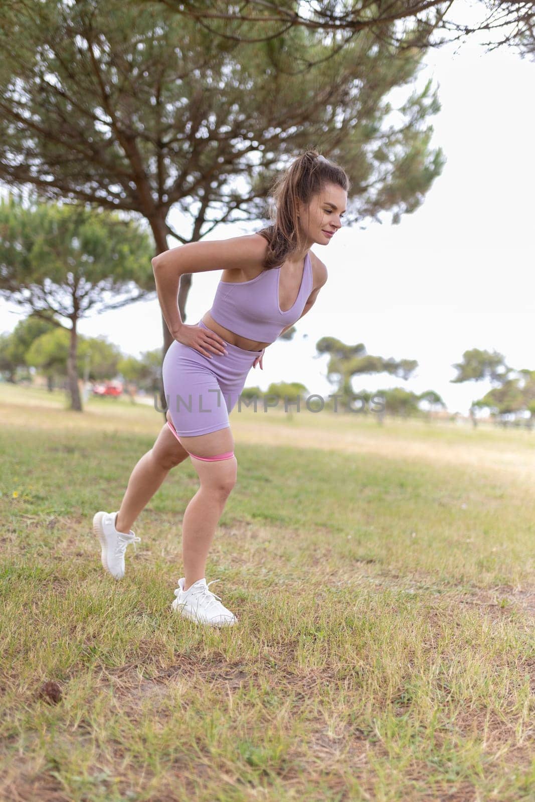 A young woman in a purple sports bra and shorts performs a leg exercise with a resistance band in a park.