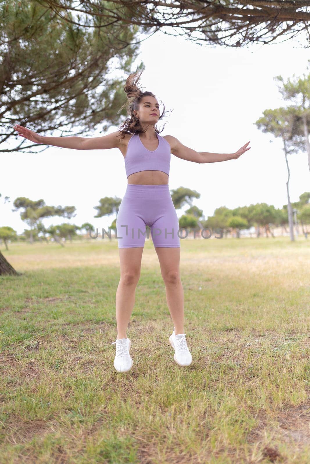 A young woman wearing a lilac sports bra and shorts jumps in the air while exercising in a park.