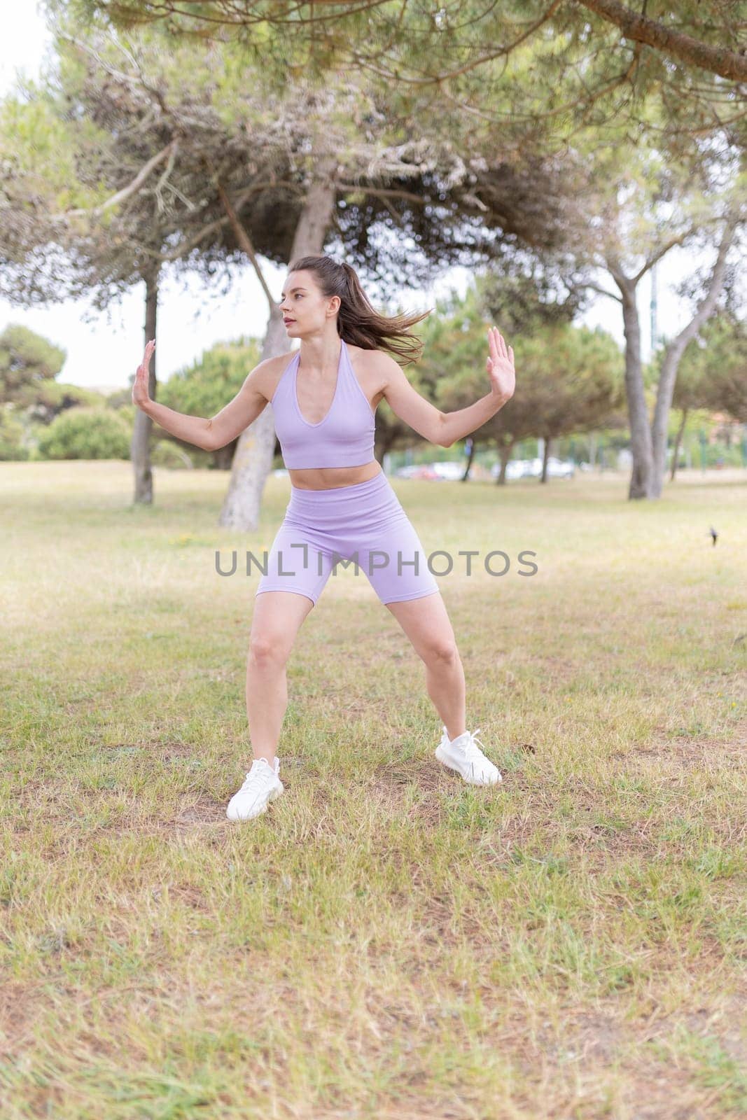 A young woman in a purple sports outfit performs a dynamic exercise in a park, surrounded by green grass and trees.