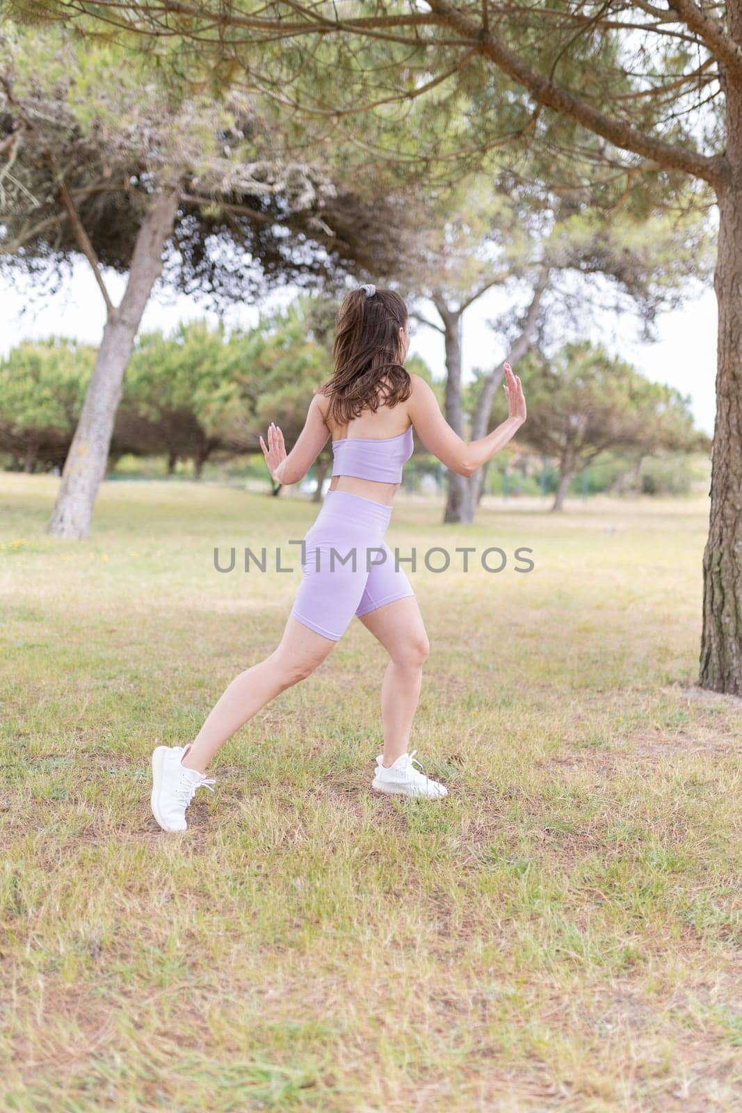 A young woman in a purple sports bra and shorts performs a dynamic stretch in a park.
