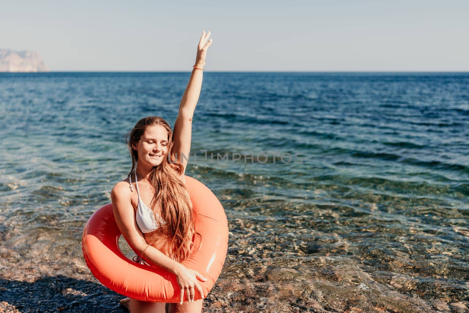 A woman is standing in the ocean with a red inflatable ring. She is smiling and waving