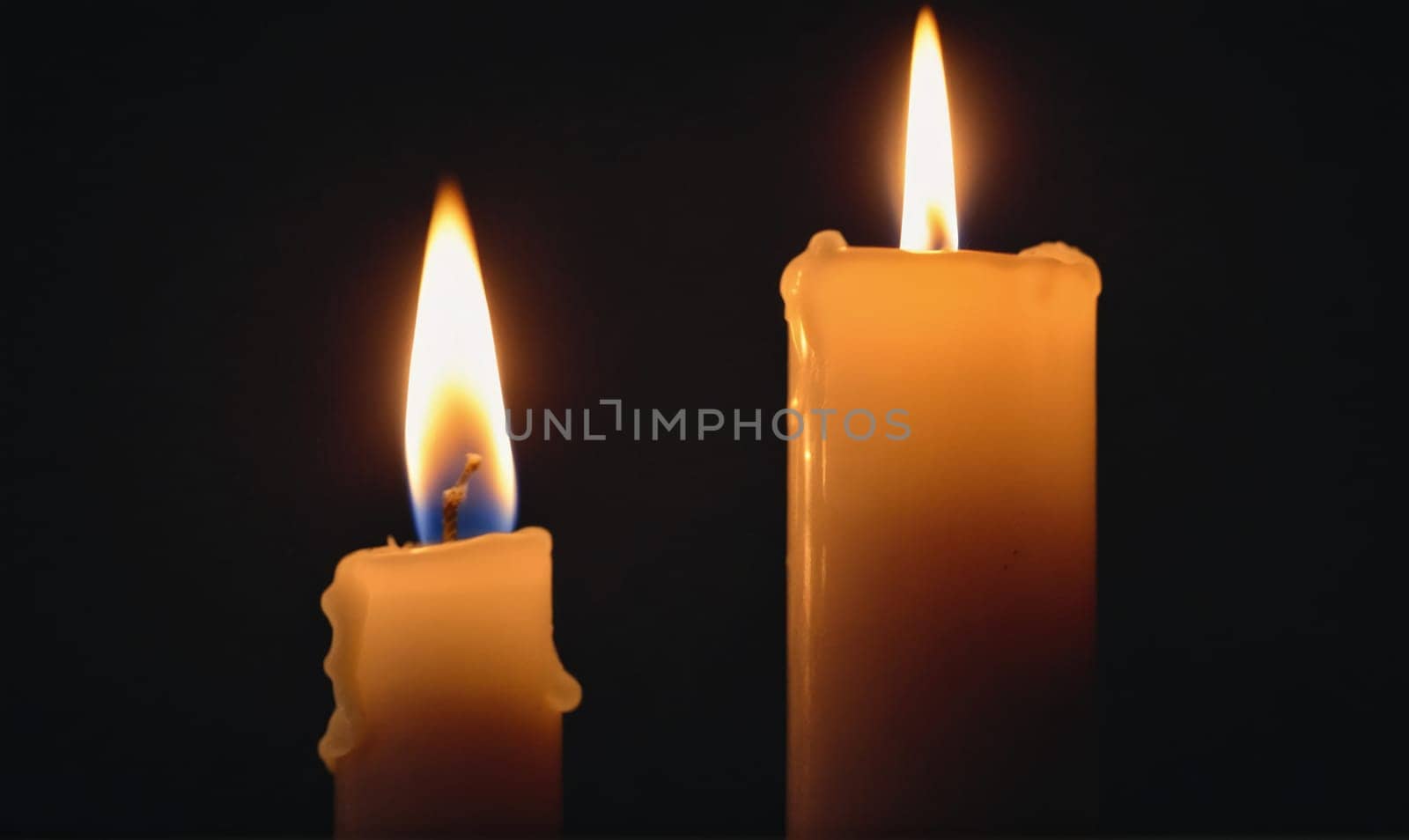 Two candles are lit in a dark room. The candle on the left is smaller and has a blue wick. The candle on the right is taller and has a white wick.