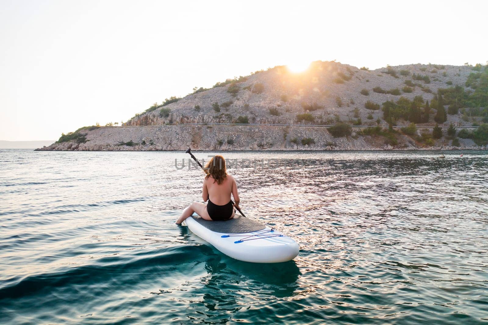 Woman paddle boarding at sunset near a rocky shoreline reflecting golden light on calm waters.