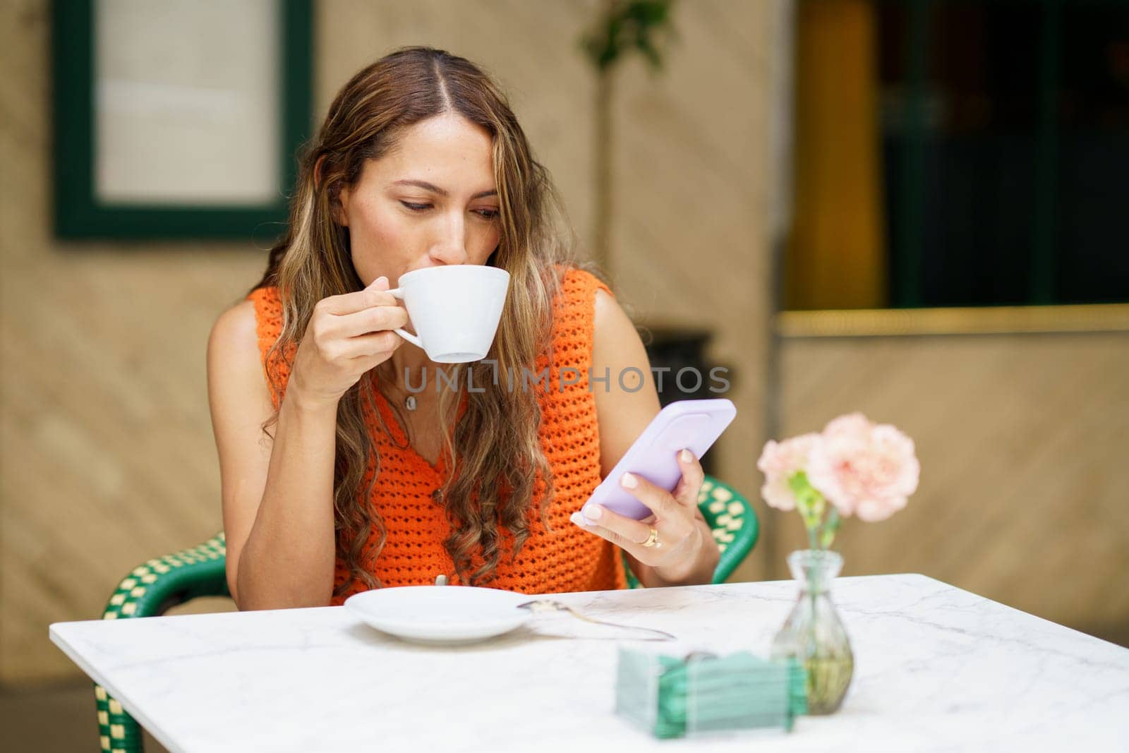 Latin woman drinking coffee while using mobile phone sitting in an outdoors cafeteria