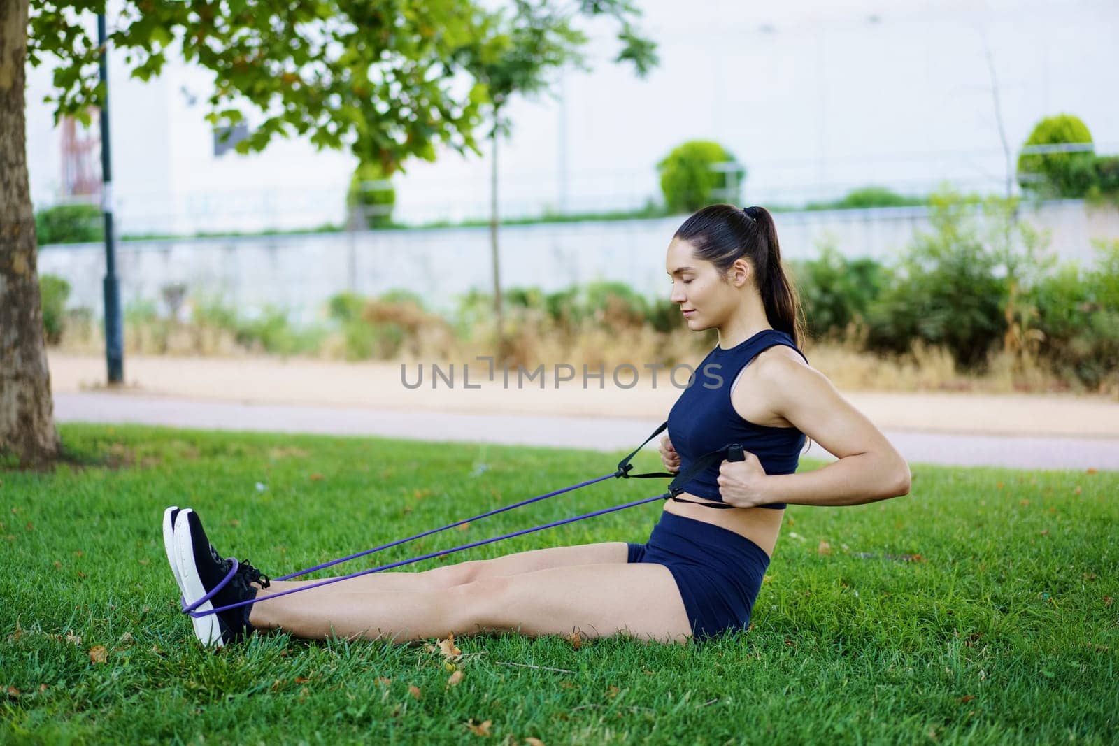 In the park, a young woman prioritizes fitness and wellness by using a resistance band for seated rows. She focuses on strengthening her back muscles as part of an outdoor workout
