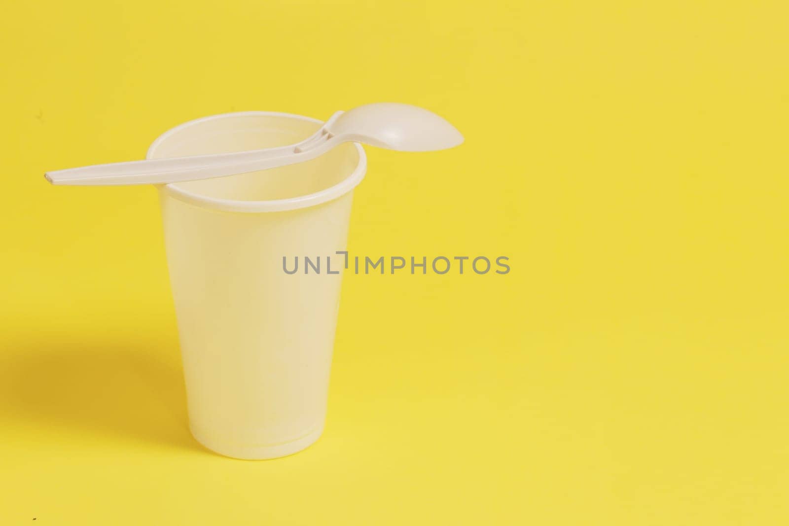 A white plastic glass and a spoon inserted into it on a yellow paper background.