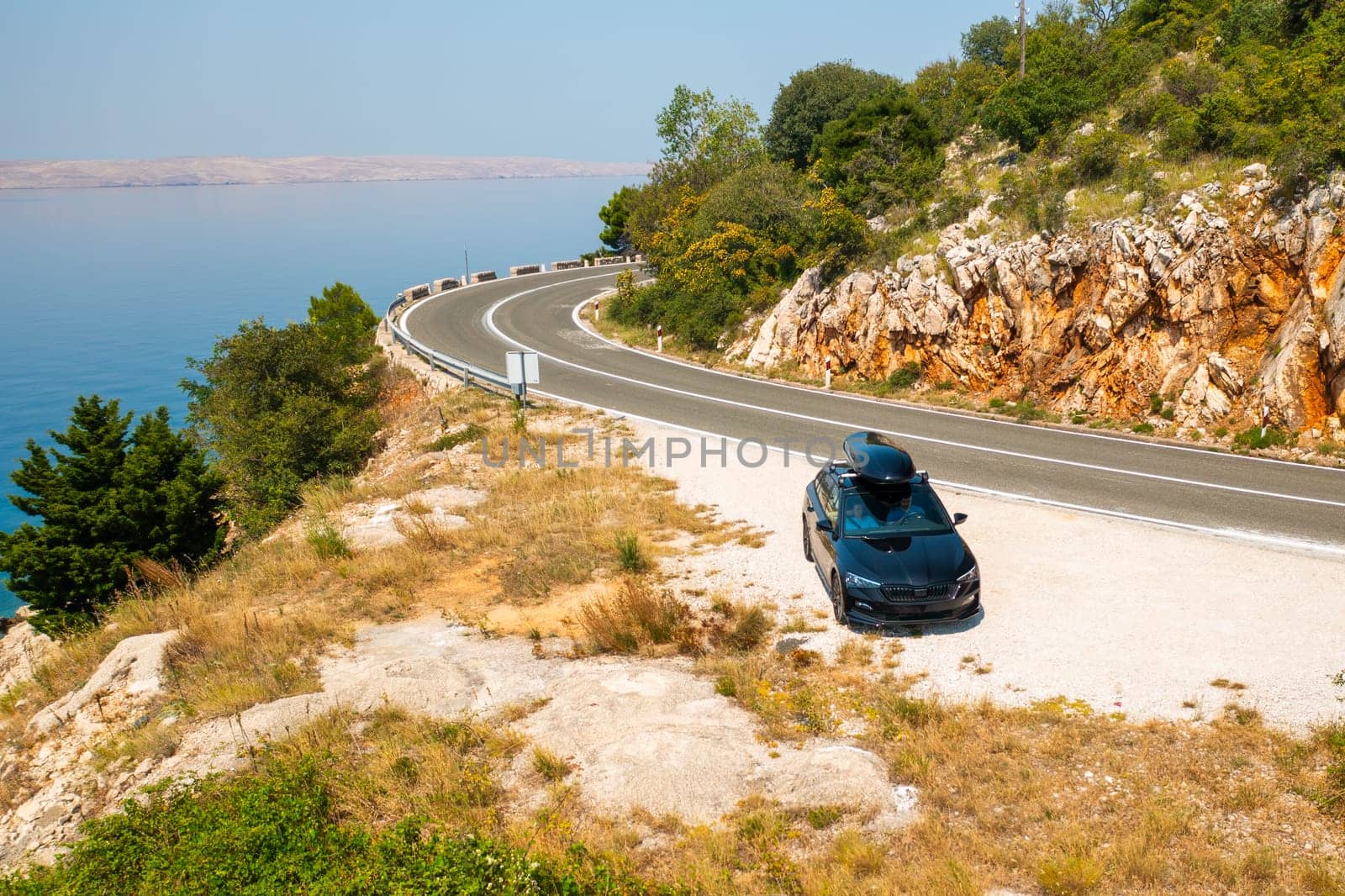 Black car with a roof box, parked at the edge of a narrow, winding road on a rugged cliff.