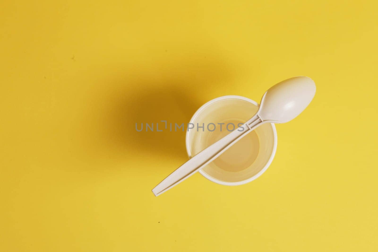A white plastic glass and a spoon lying on it on a yellow paper background.
