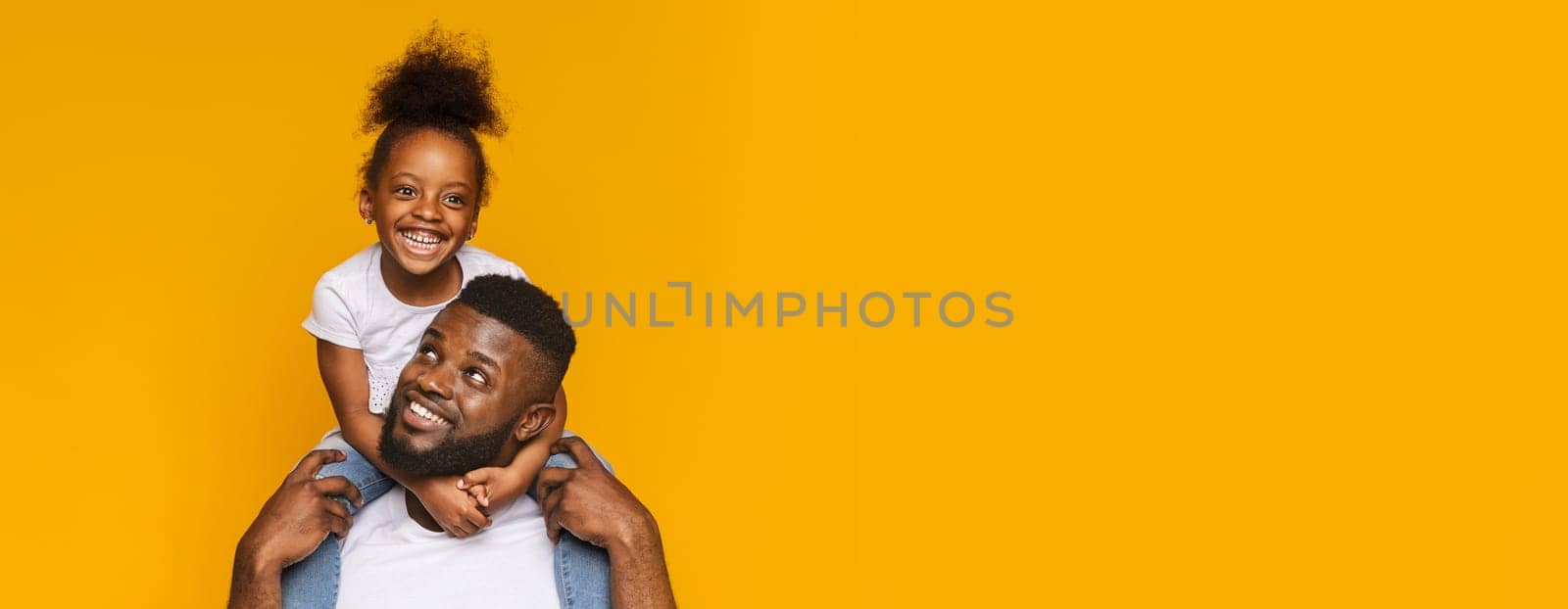 African American father happily carries his daughter on his shoulders, both beaming with joy. They are set against a vibrant yellow backdrop that enhances their cheerful expressions, copy space