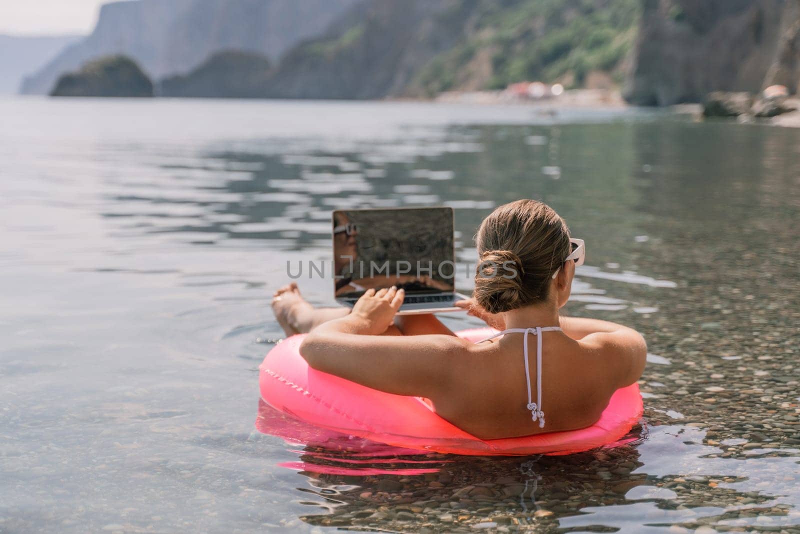 A woman is sitting on a pink inflatable raft in the water, using a laptop. Concept of relaxation and leisure, as the woman is enjoying her time in the water while working on her laptop