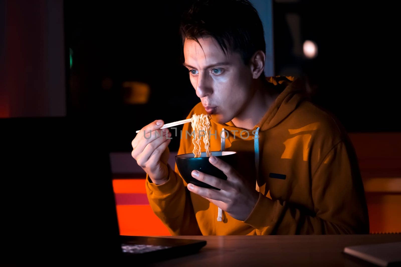 A young man in a yellow casual hoodie eats noodles with sticks and watches a movie at night on a laptop at home, a man works, studies and has a snack.