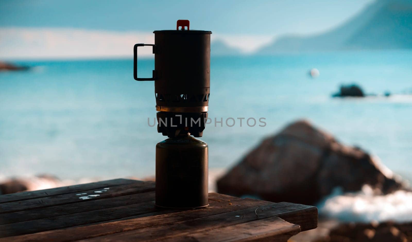 Camping burner stands on a table against the backdrop of a picturesque landscape with a beach and sea at sunset, camping equipment for quick cooking without the use of a fire with wood.