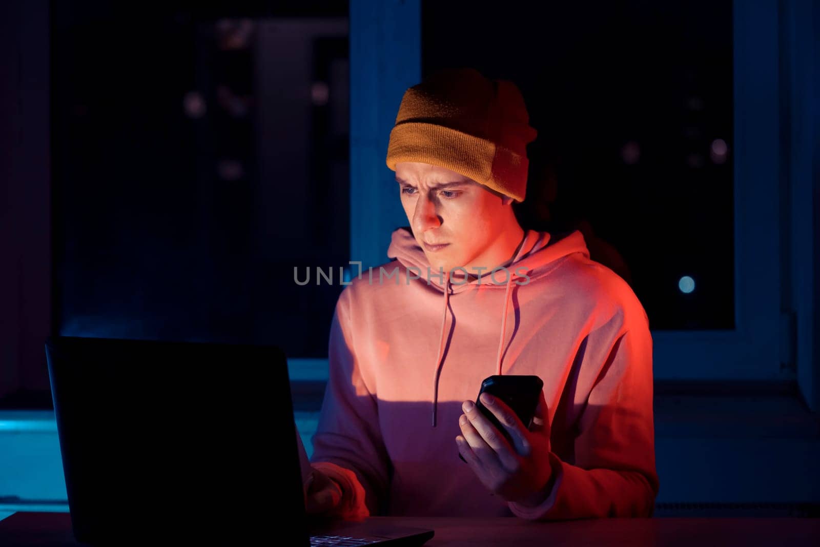 A young man works late at night at a laptop at home and talks on the phone, solves business issues, analyzes files, freelancer communicates online.