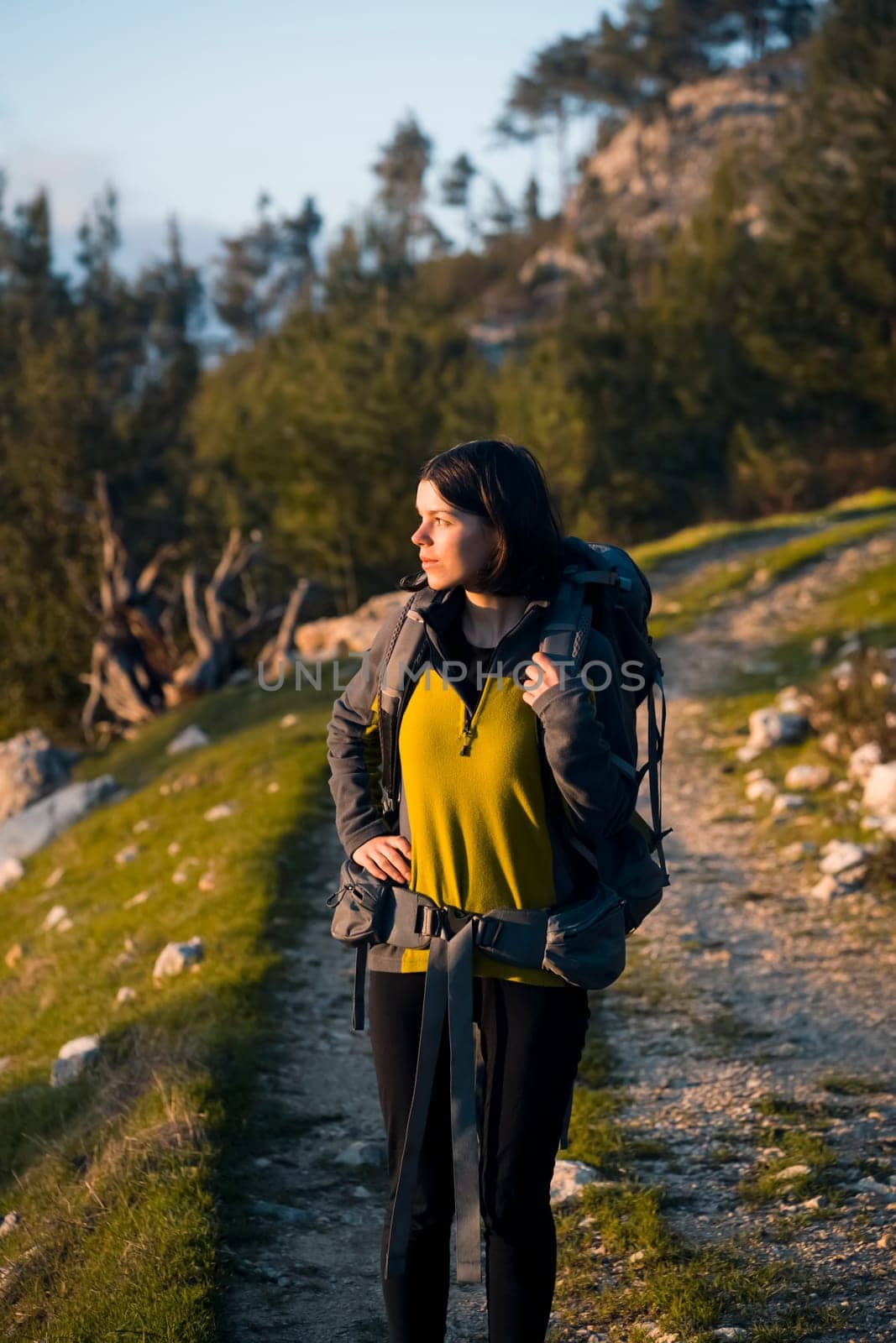 A young girl with a backpack and travel equipment looks at the amazing sunset in the mountains while she hikes along the trail. A traveler walks through the national park.