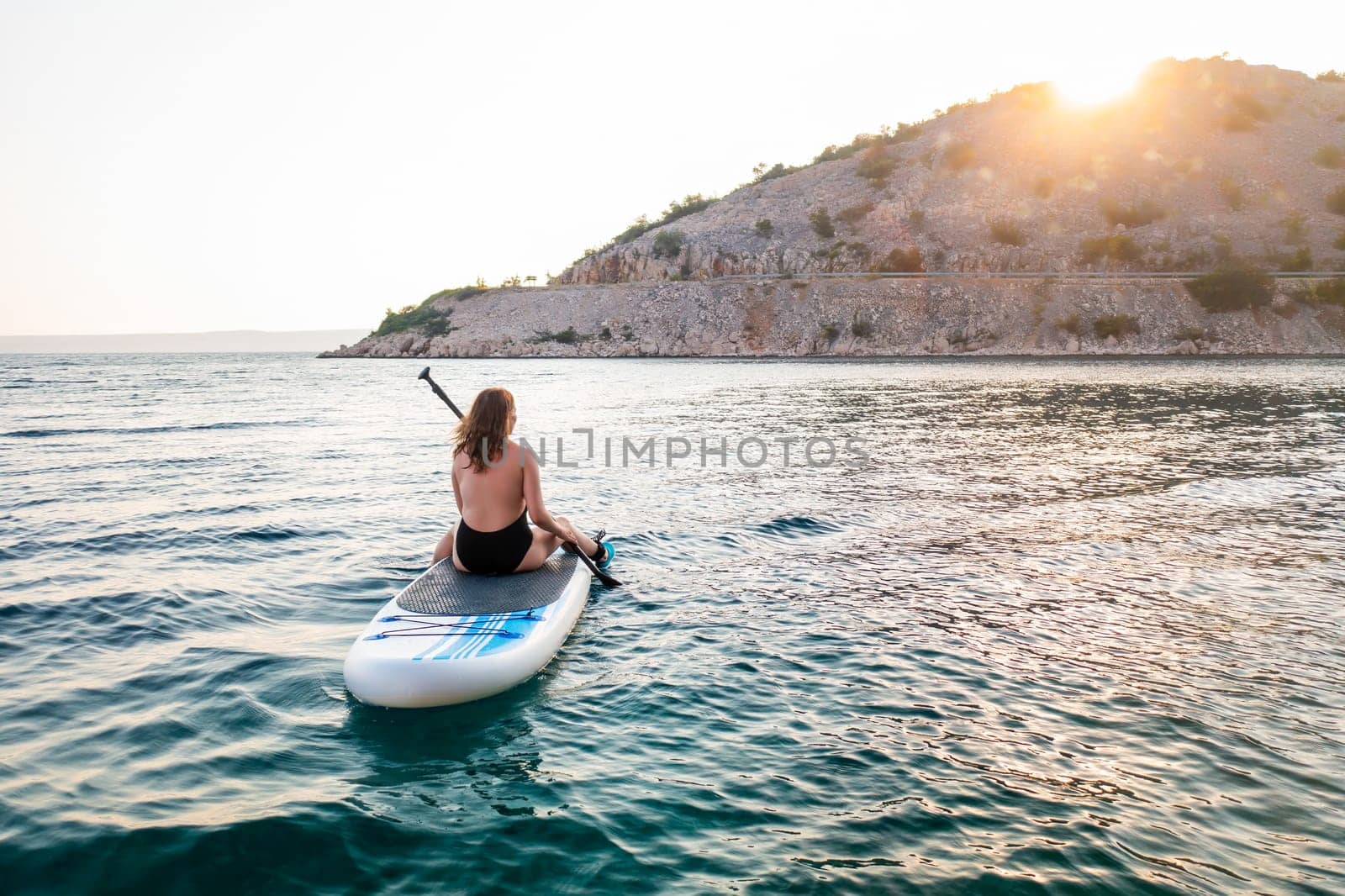 Woman paddle boarding on serene waters during sunset near a rocky shoreline.