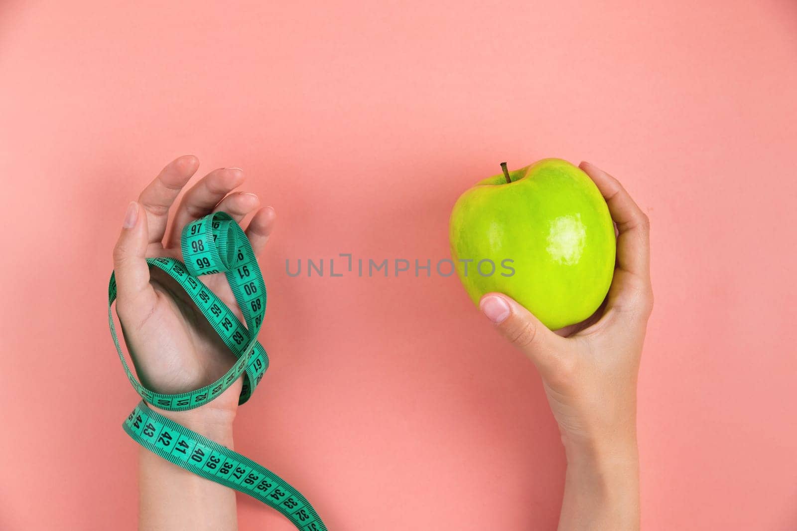 woman's one hand holds a green apple and the other hand holds a measuring tape, on a pink background. Weight loss and diet concept by yanik88