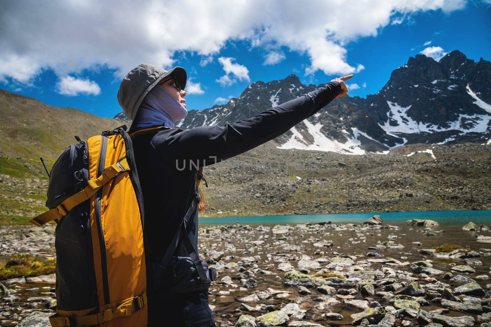 Close up of young Caucasian female traveler standing, showing destination mountains, pointing finger to right outdoors. Snowy mountains with a lake, a girl with a backpack on a sunny day.