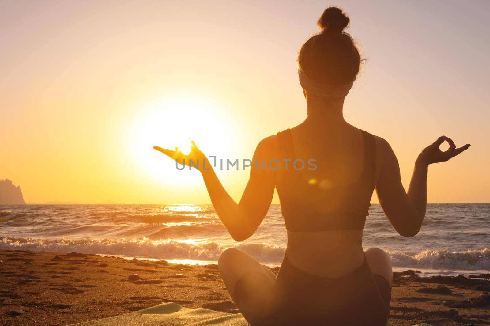 A young caucasian woman in sportswear sits on the seashore in the early morning or at sunset in a lotus position with mudras of bragost in her hands. Silhouette back view.