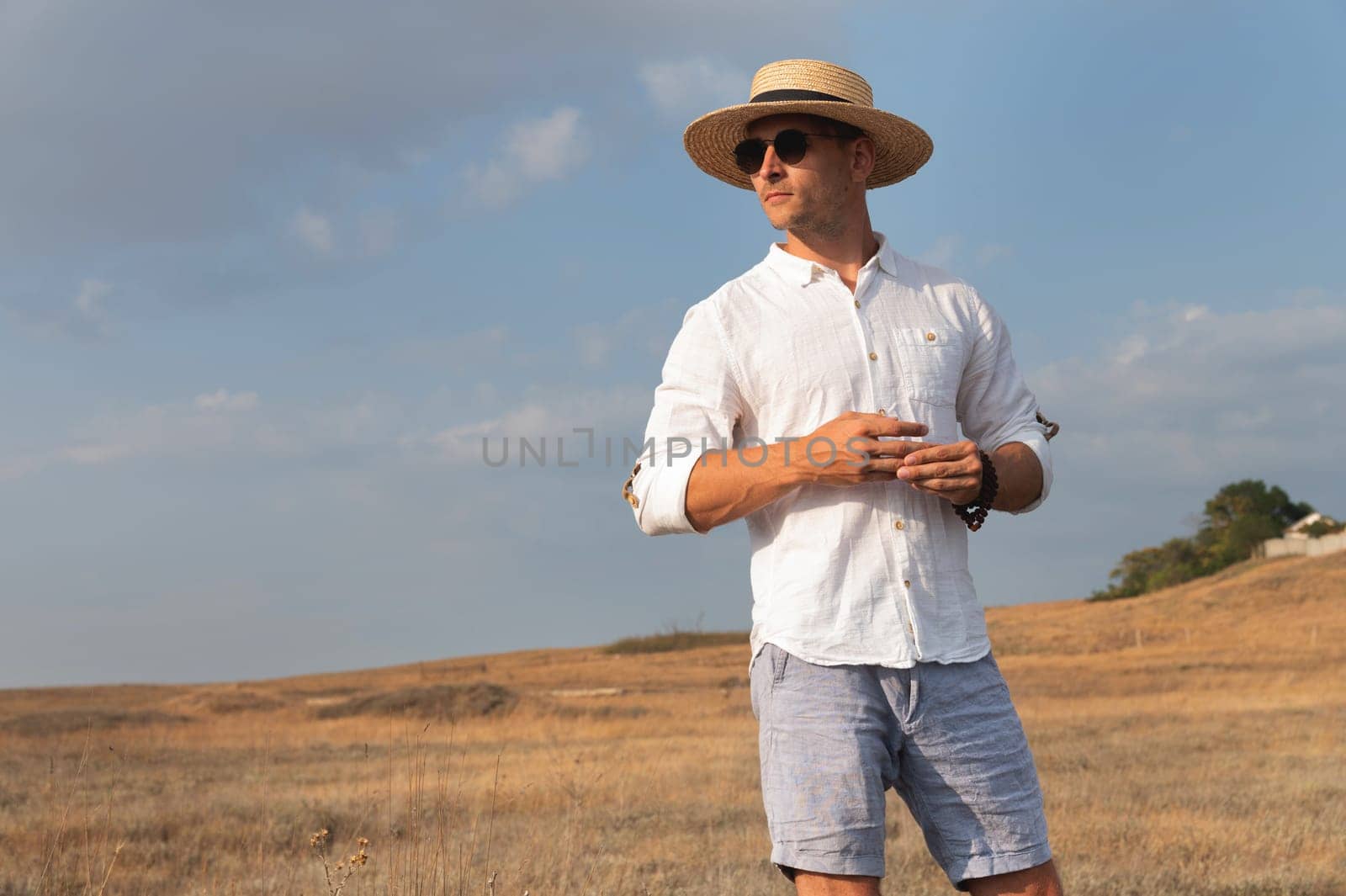 Young sexy muscular man posing outdoors looking away, wearing a straw hat and sunglasses. coastal field.