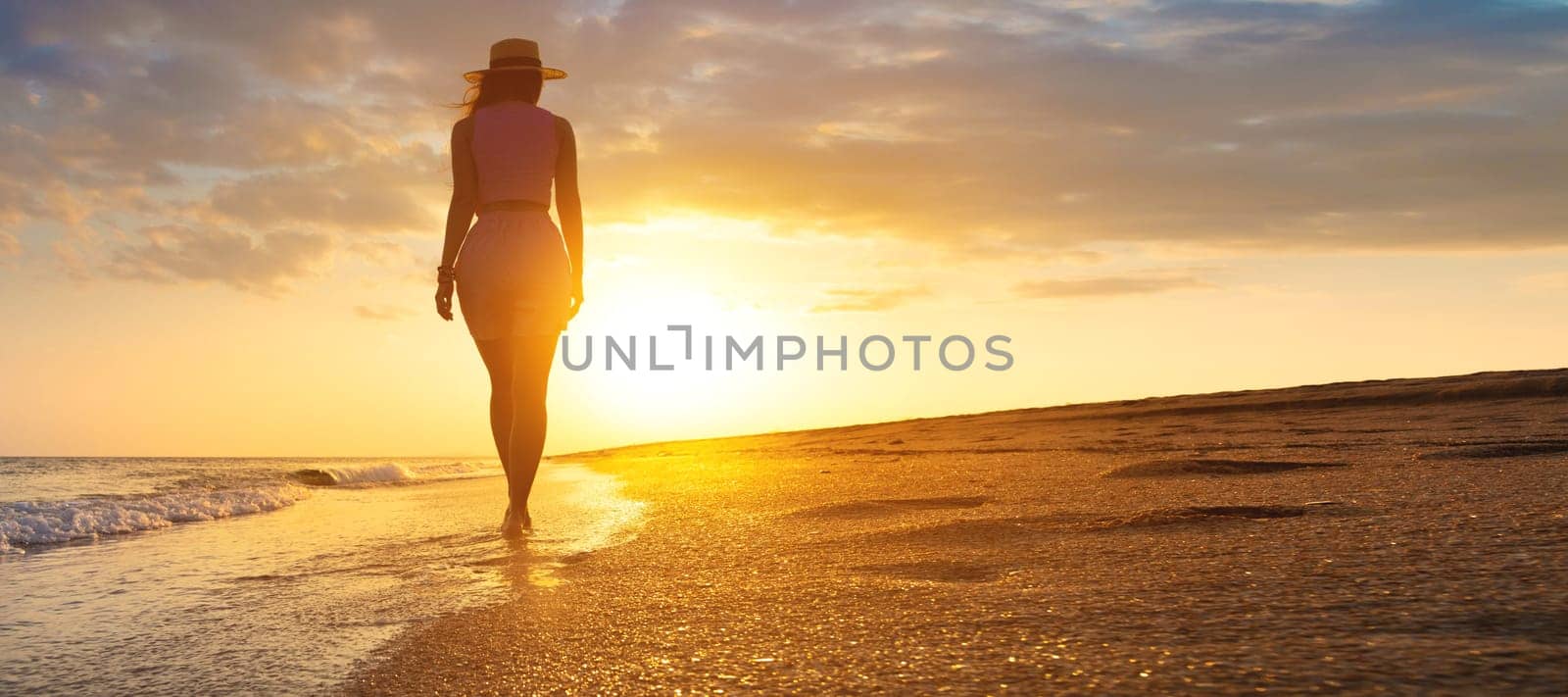 Photo of a young stunning woman walking along the seashore. Fresh sea breeze, a girl watches the sunset and walks towards the sun's rays.