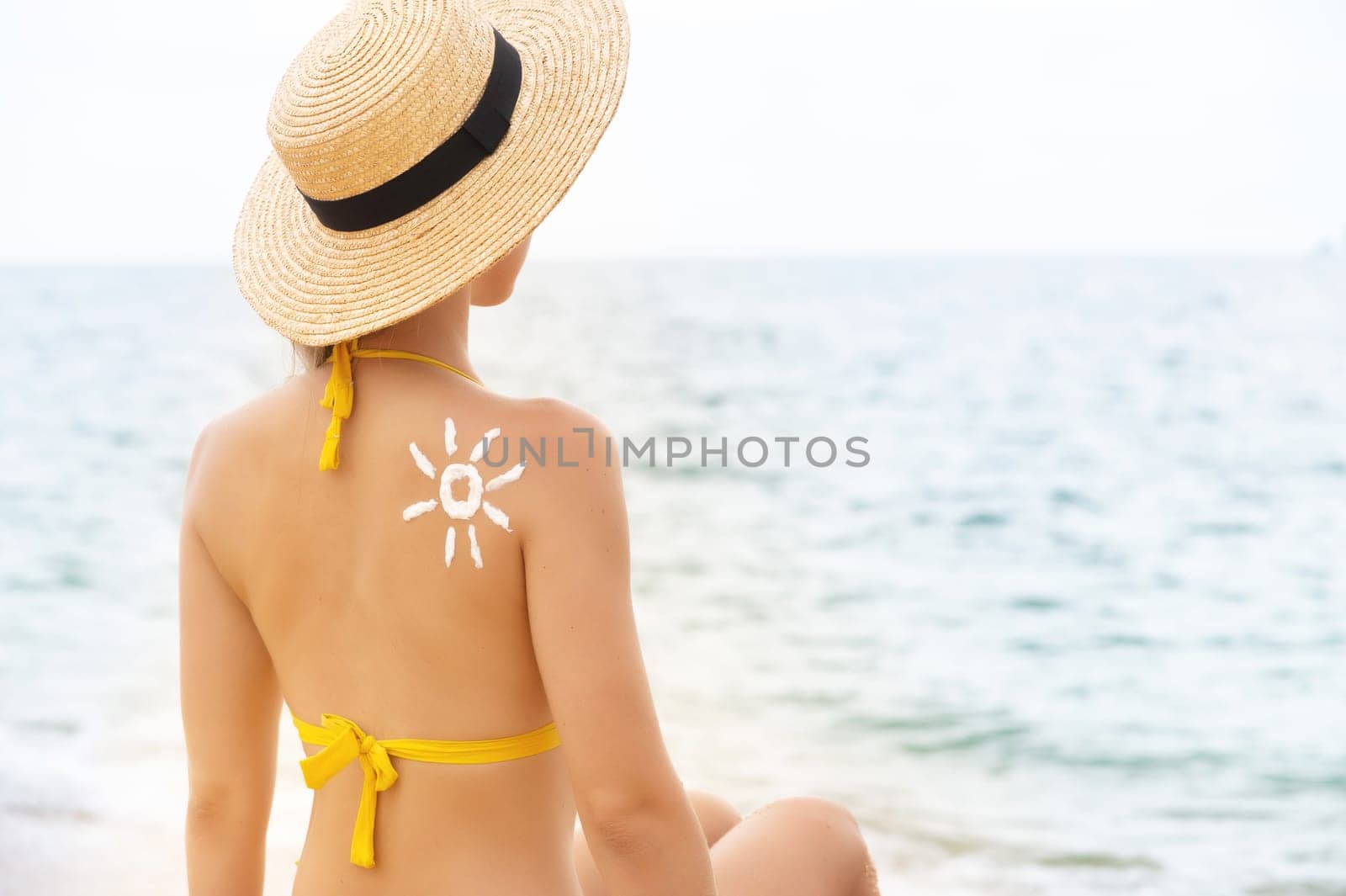 Caucasian girl with sun-shaped sunscreen on her shoulder on a sandy beach. Beautiful young woman sitting by the sea on vacation, sunbathing.