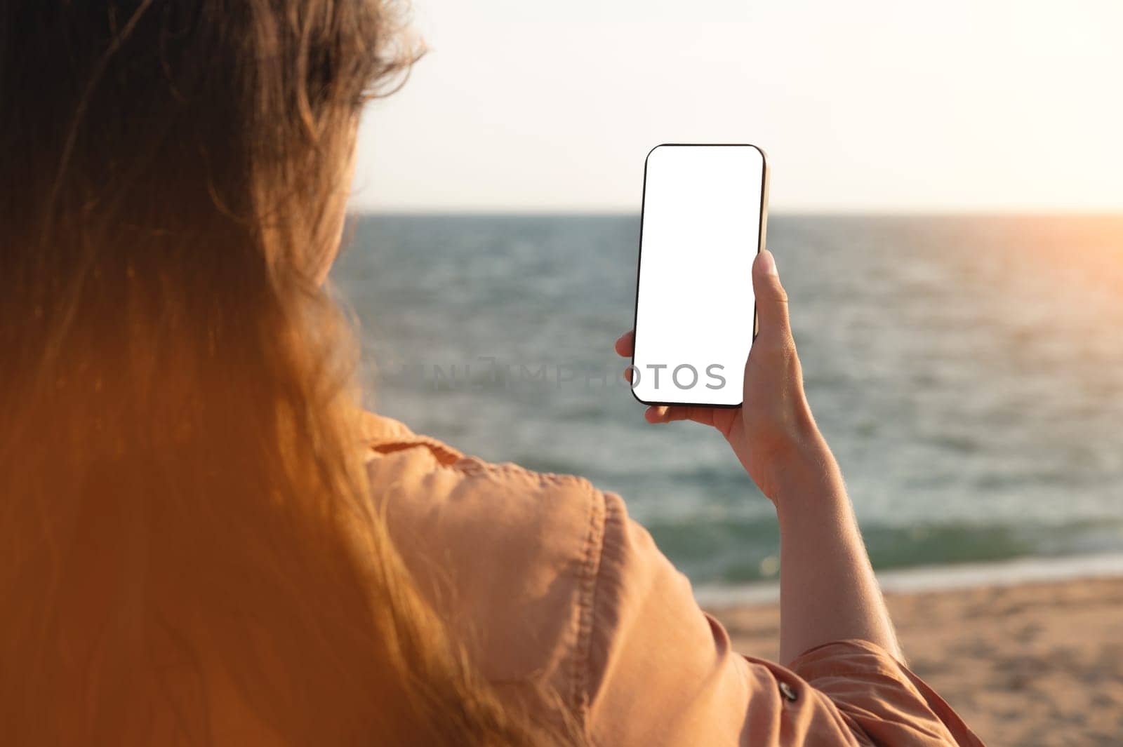 Hand holding smartphone on the beach, mobile blank screen mockup. Screen to place advertisements. Summer vacation concept, woman's hand with a phone on the background of the sea by yanik88