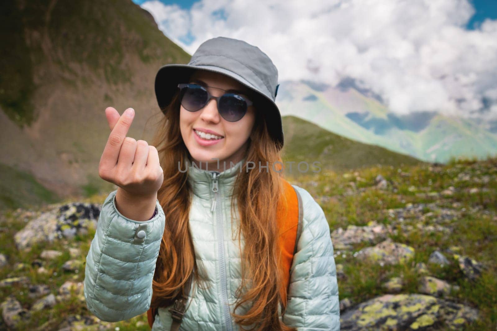 Attractive young Caucasian woman feels happy, shows a heart gesture, expresses tender feelings, against the backdrop of mountains and cloudy sky. Caring for people concept by yanik88