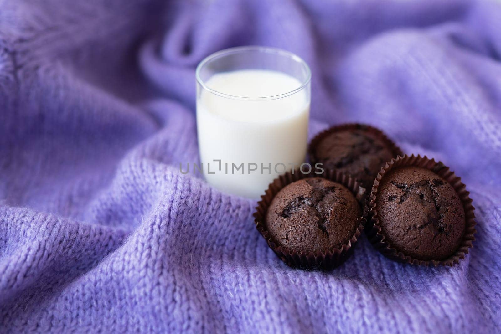 Chocolate cupcakes, along with a glass of milk, stand on a purple knitted fabric. Close-up, place for an inscription