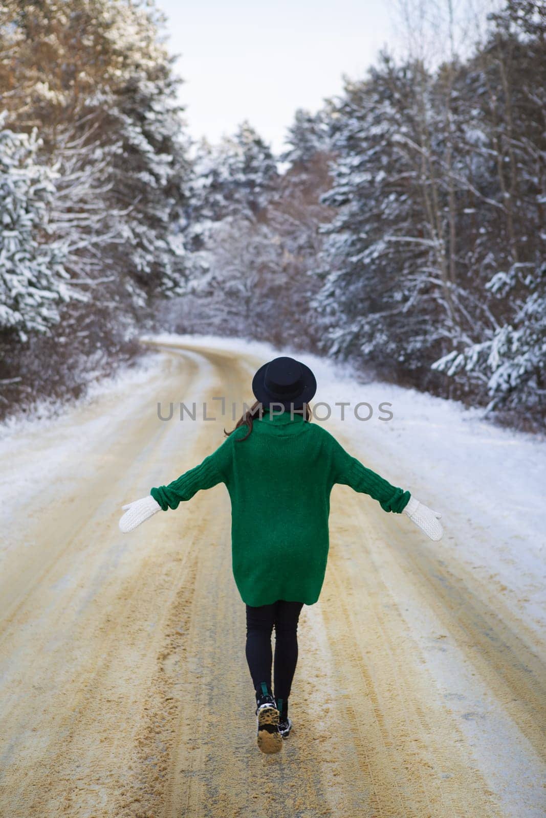 A young girl in a green sweater and hat walks in the middle of a snowy road in a thick pine forest. Freezing day