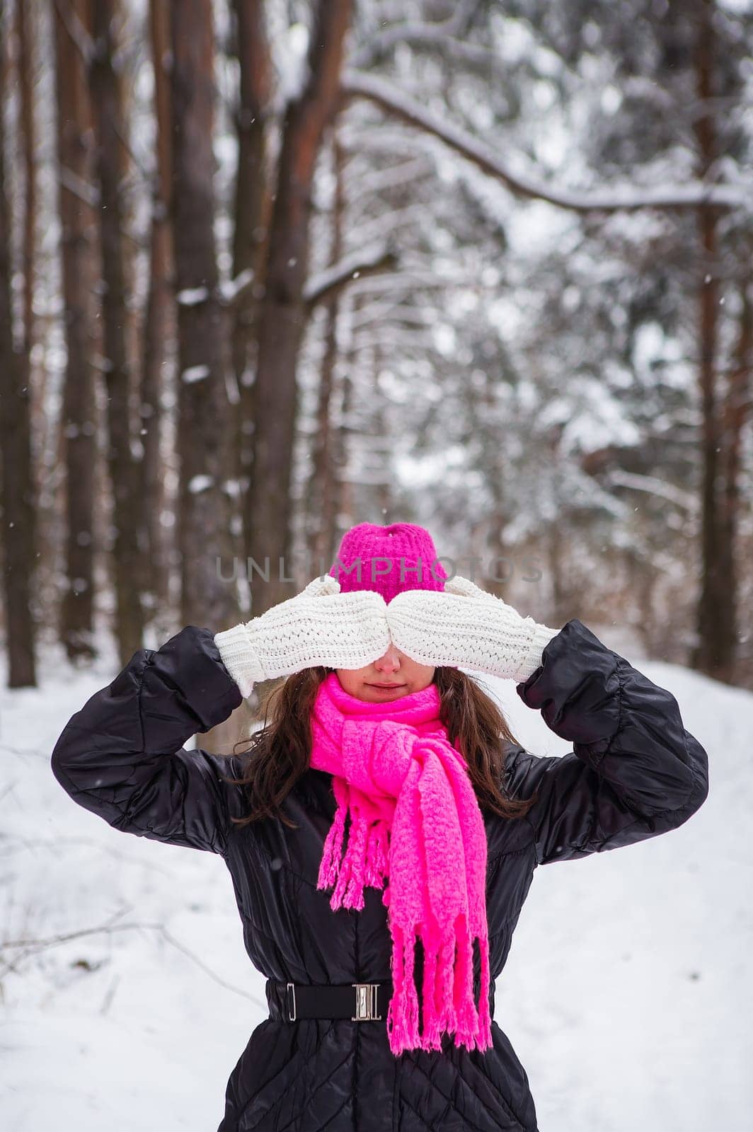 A girl in a pink scarf and hat stands in the middle of a pine forest and covers her face with white mittens. Beautiful pine forest on a frosty day
