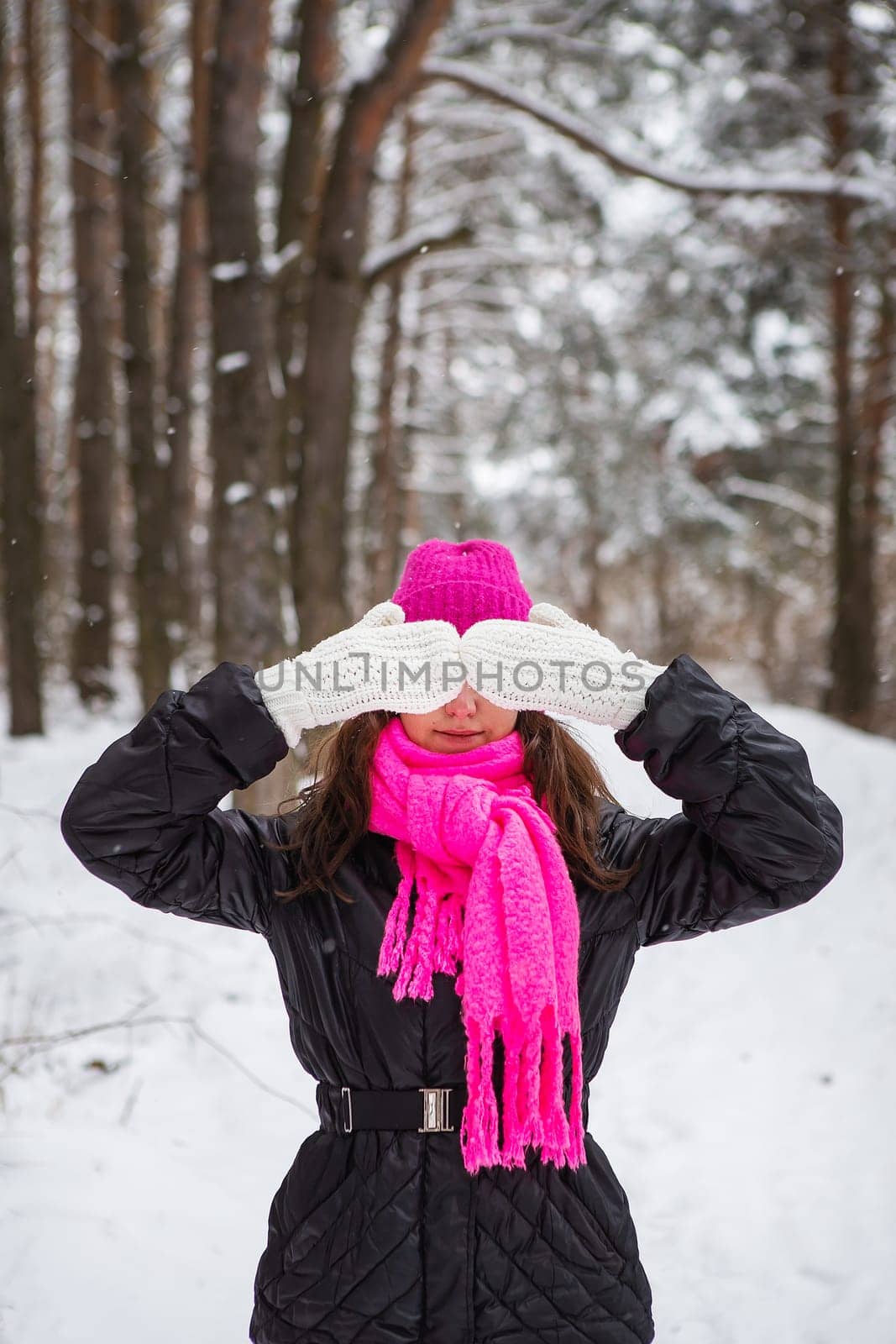 A girl in a pink scarf and hat stands in the middle of a pine forest and covers her face with white mittens. Beautiful pine forest on a frosty day. by sfinks