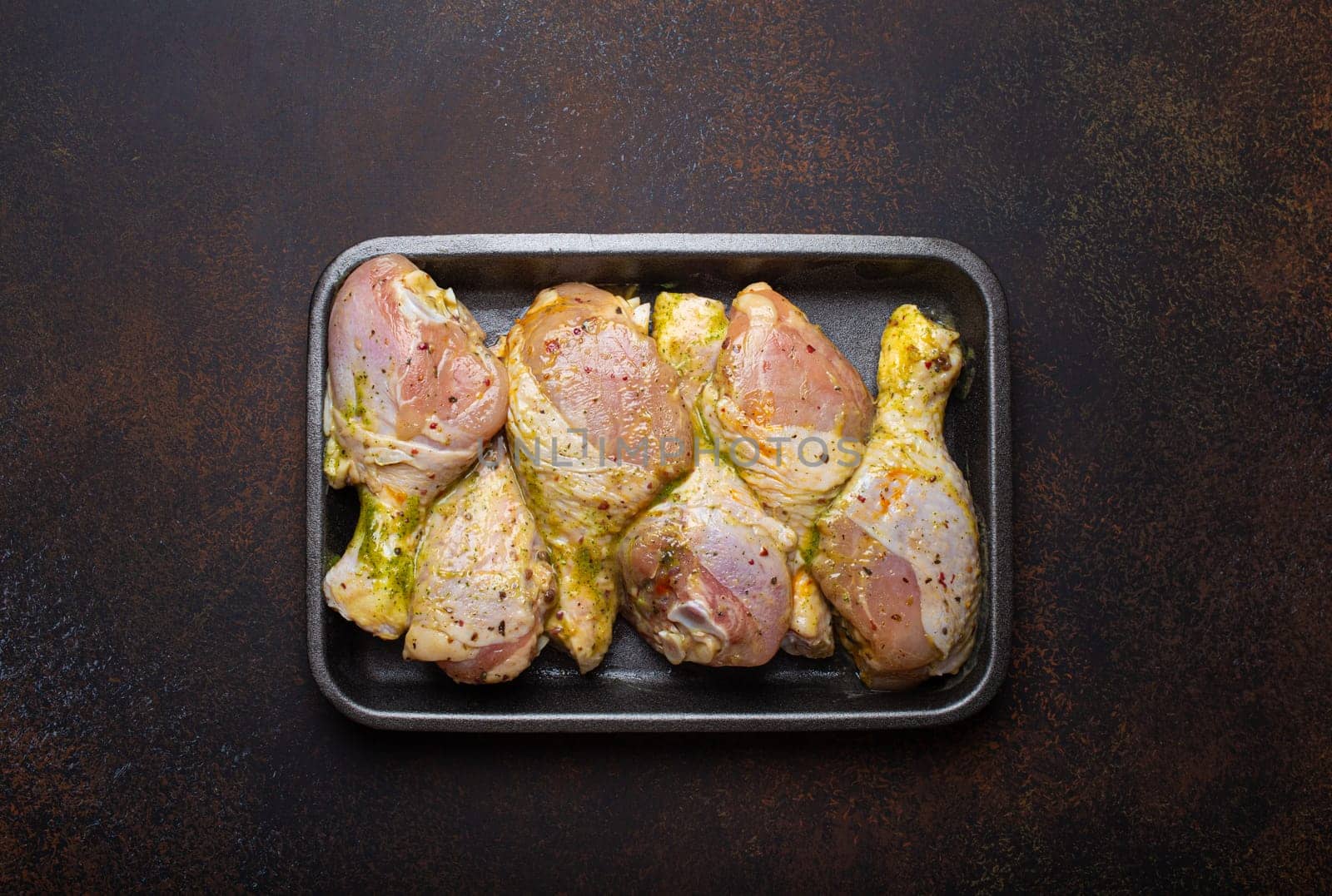 Raw uncooked chicken legs in green marinade with seasonings in black plastic container top view on dark rustic background. Preparing healthy meal with marinated chicken drumsticks by its_al_dente