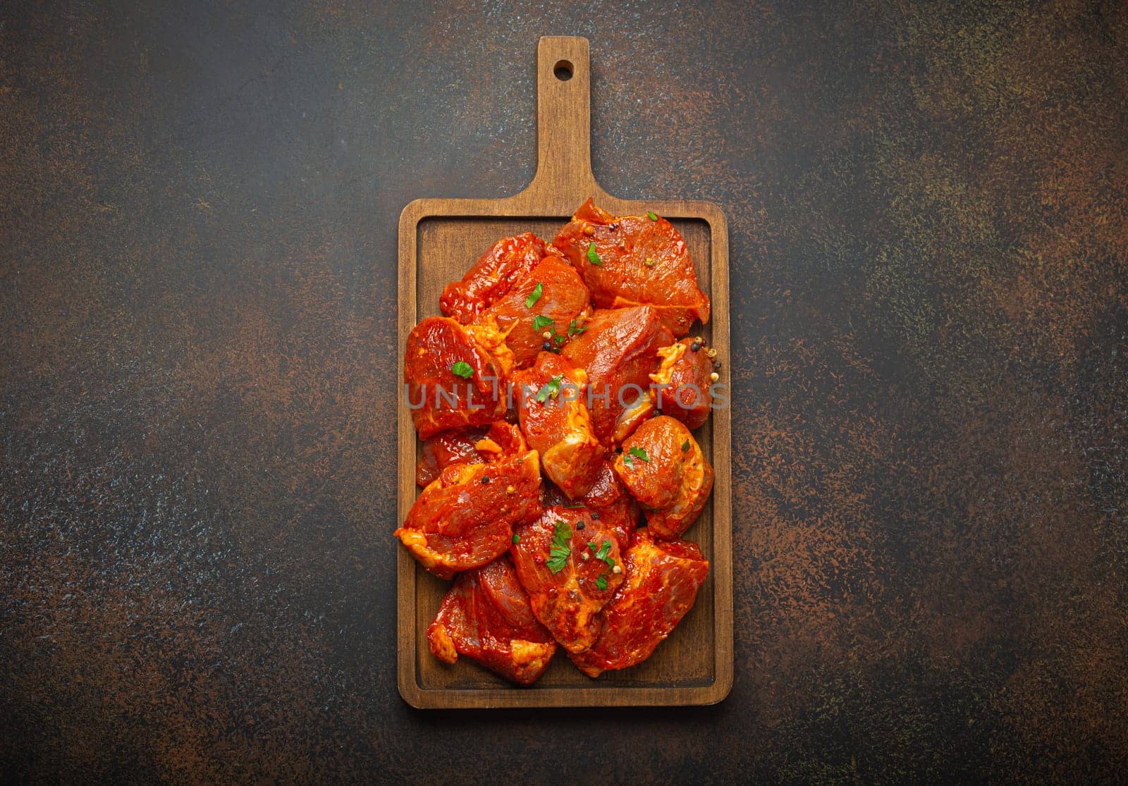 Raw uncooked chopped pieces of pork marinated with seasonings and parsley on wooden cutting board top view on dark rustic background. Cooking meal with marinated pork fillet pieces by its_al_dente