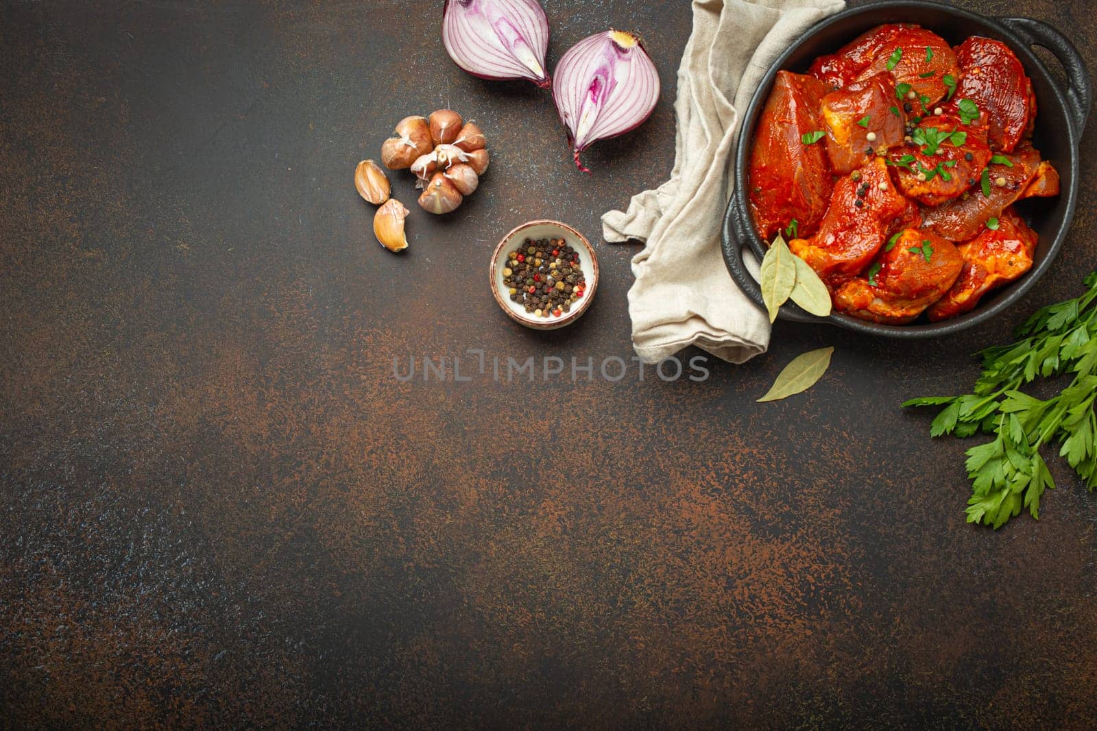 Cooking hearty meat stew. Raw uncooked chopped pieces of meat marinated with seasonings and parsley in black casserole dish top view, ingredients on dark rustic background. Space for text.