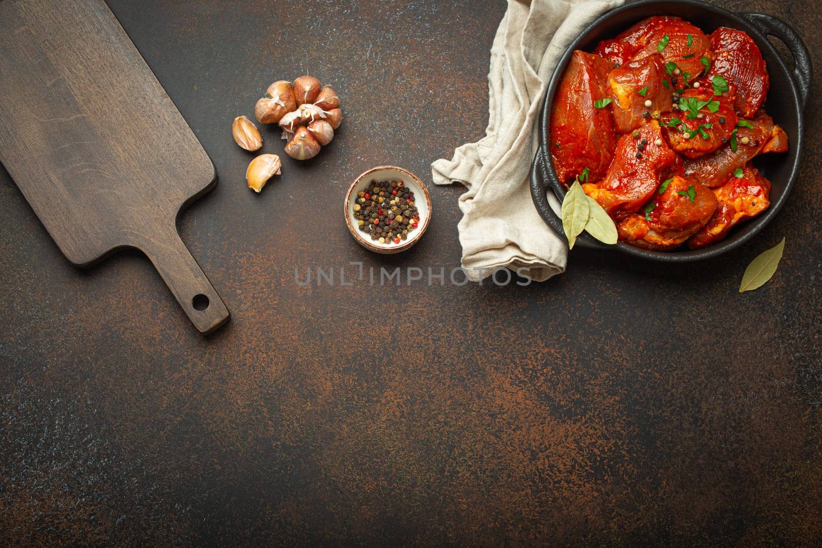 Cooking meat stew. Raw uncooked chopped pieces of meat marinated with seasonings and parsley in black casserole dish top view, cutting board, ingredients on dark rustic background. Space for text.