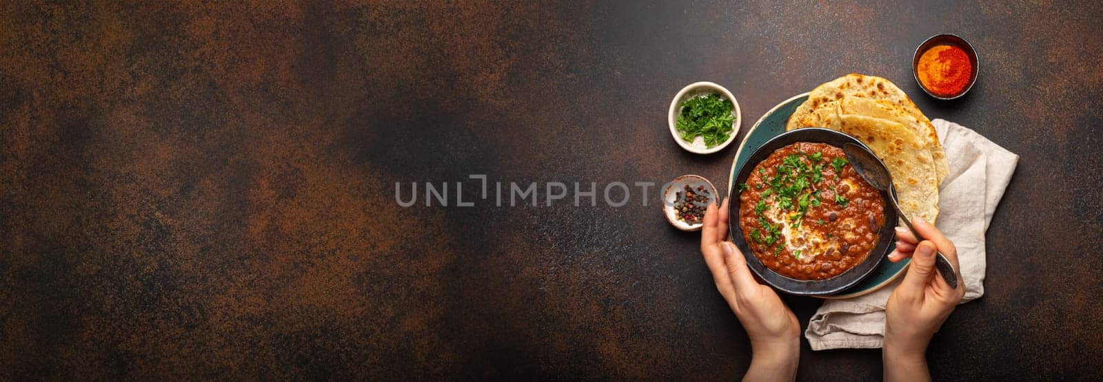Female hands holding a bowl, eating traditional Indian Punjabi dish Dal makhani with lentils and beans served with naan flat bread, fresh cilantro on brown concrete rustic table top view. Copy space by its_al_dente