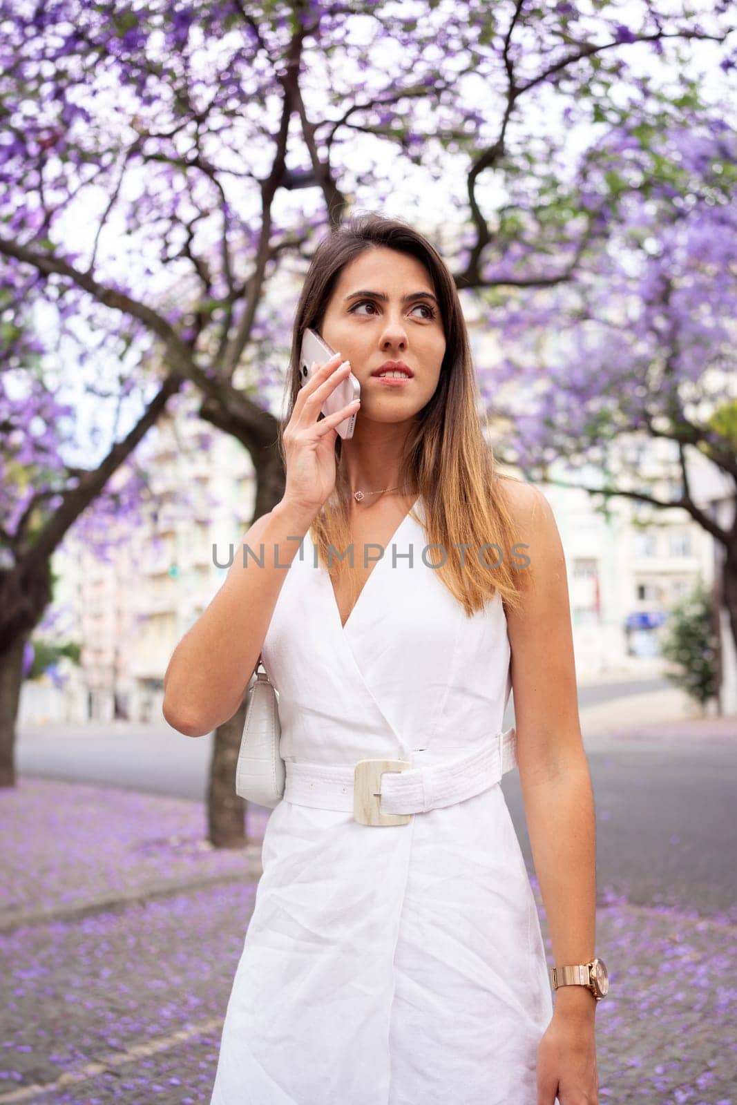 Woman in white dress uses cellphone outdoors. Female making call in city park. Fashionable female wearing white summer dress stands outdoors with violet Jacaranda trees in background.