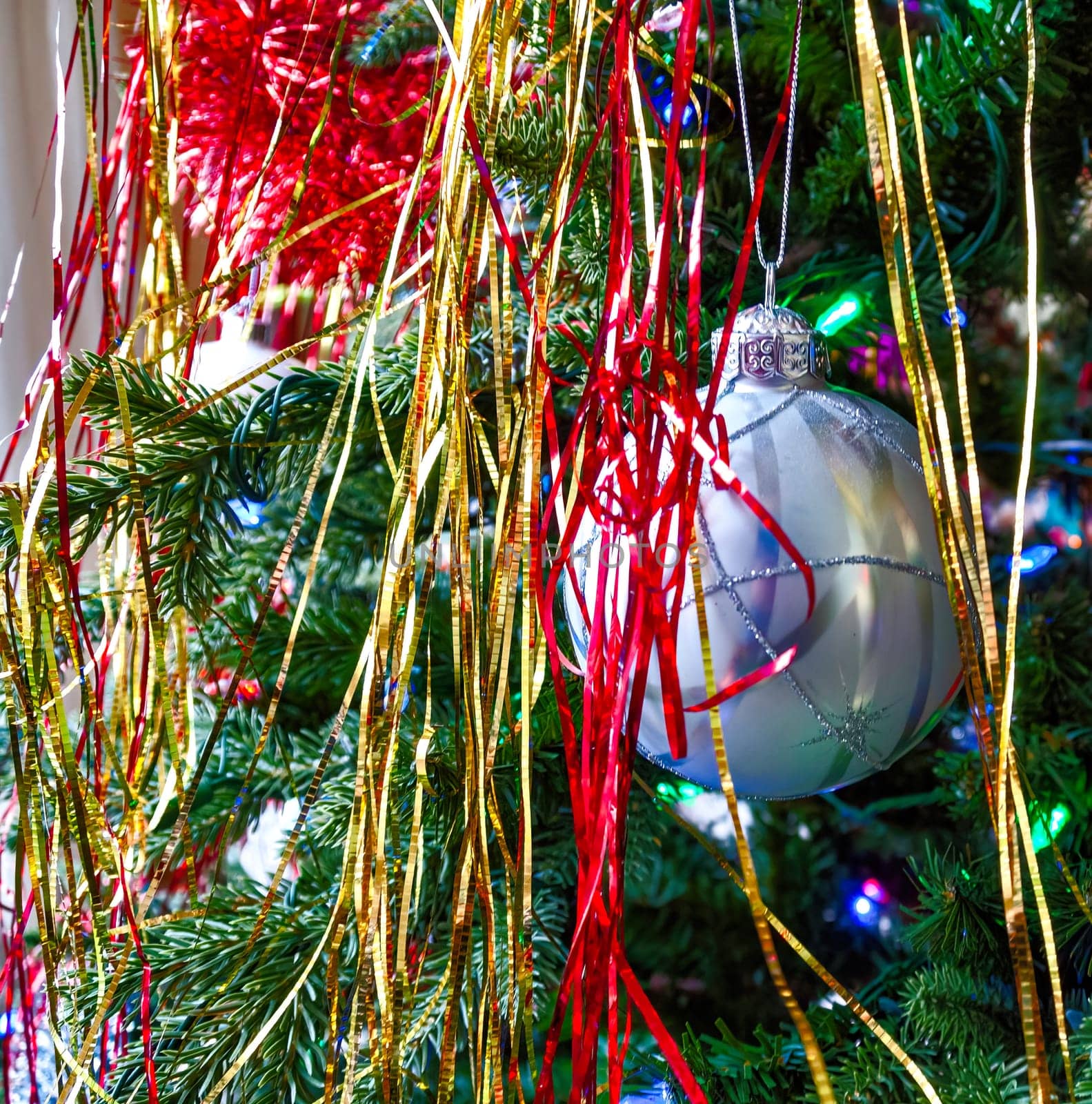Silver bauble with silver straps on chistmas tree among decorations. Closeup look of xmas ball on the tree