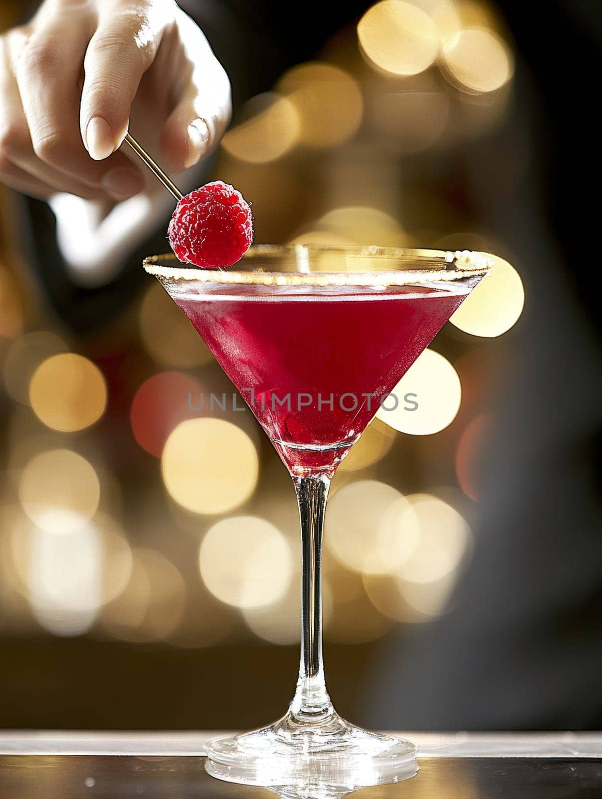 Elegant red cocktail in a martini glass with a raspberry garnish being delicately placed by hand, set against a blurred background of warm lights.