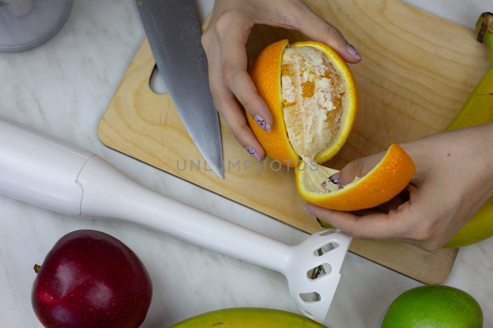 Female hands peeling orange on cutting board in kitchen while preparing sports drink with blender. Healthy eating concept with vitamins in fruits