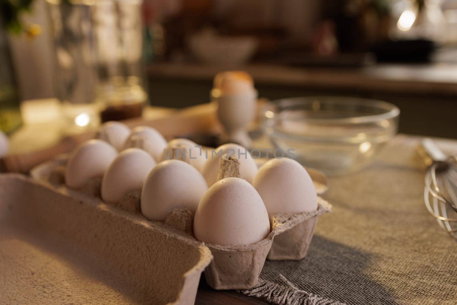 A close-up of a carton of white eggs on a kitchen countertop, with a blurred background showing a person and kitchen utensils