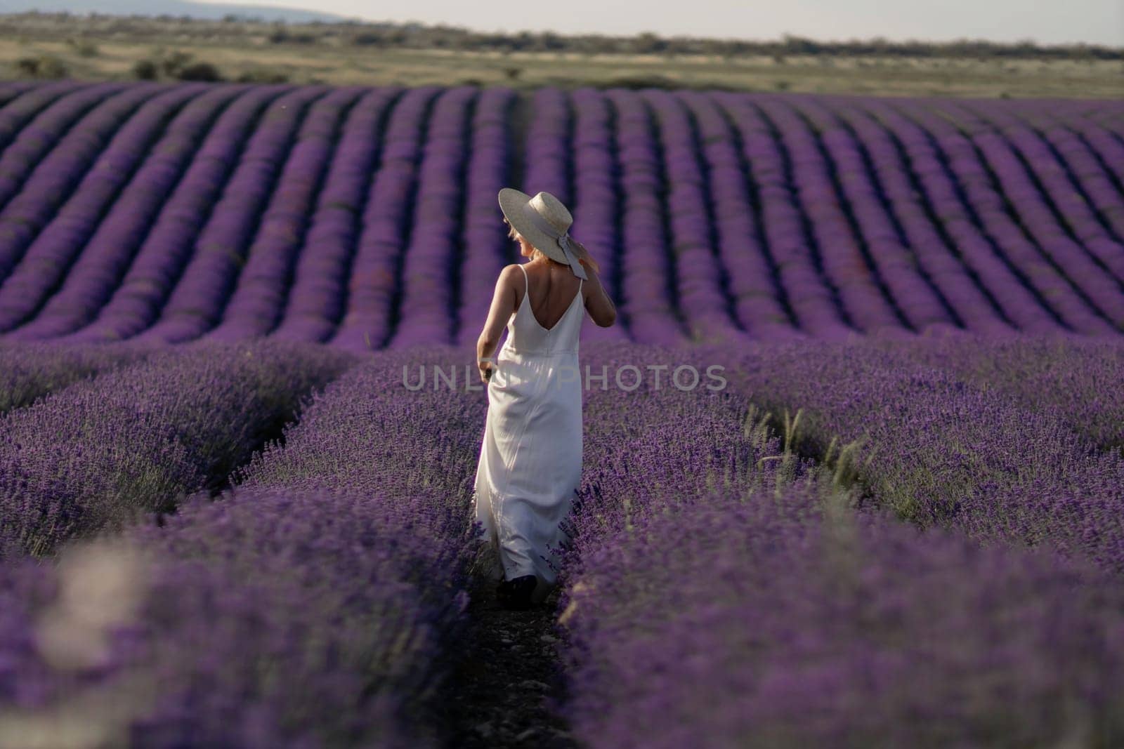 A woman is walking through a field of lavender. The field is vast and the flowers are purple. The woman is wearing a white dress and a straw hat
