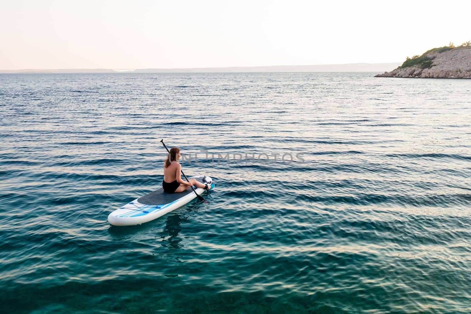 A woman paddle boarding on tranquil waters near a rocky shoreline at sunset.
