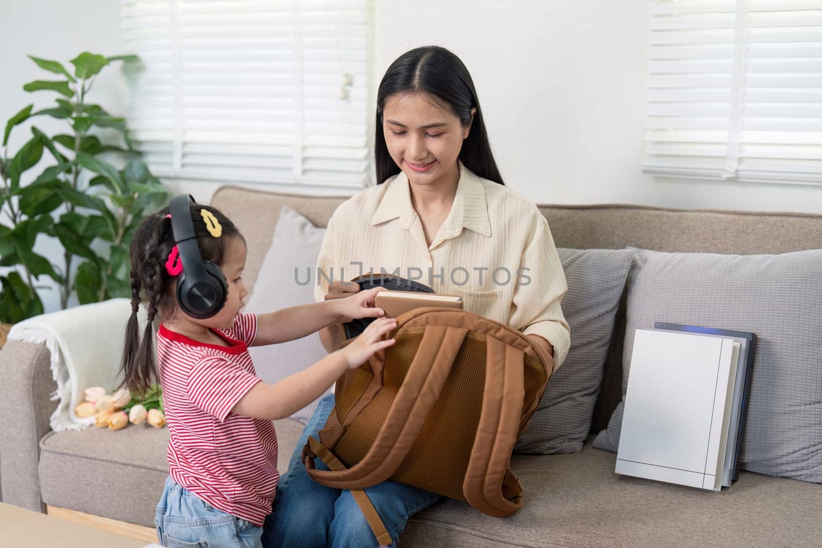 Little girl listens with headphones while her mother organizes school items in a home setting, highlighting back-to-school preparation