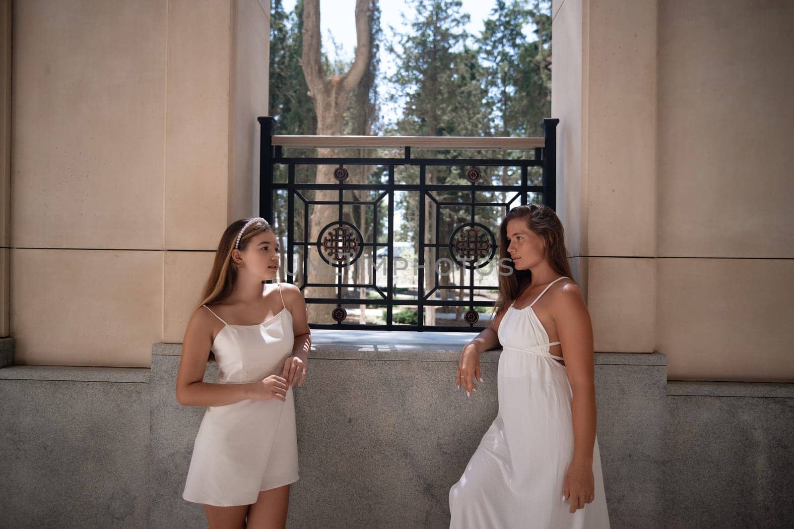 Two women in white dresses stand by a window, one holding a flower, while the other leans against the wall.