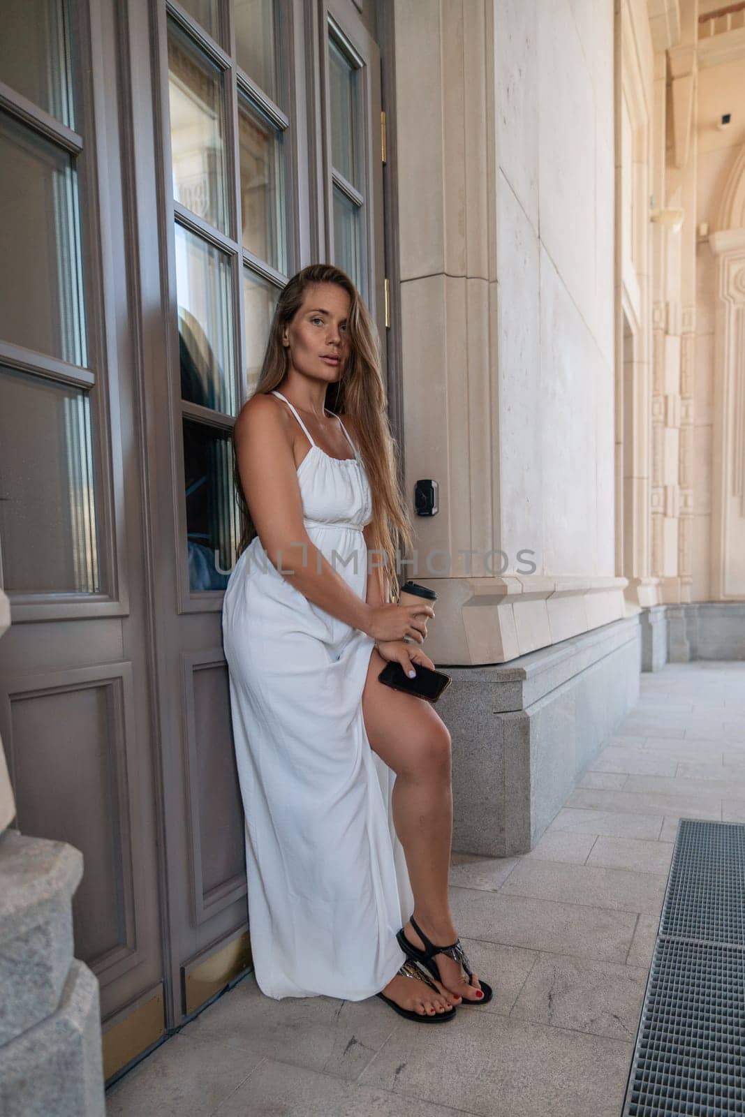 A woman in a white dress stands beside a wooden door, her right hand resting on the door frame. Her left hand holds a black purse. She is looking at the camera.