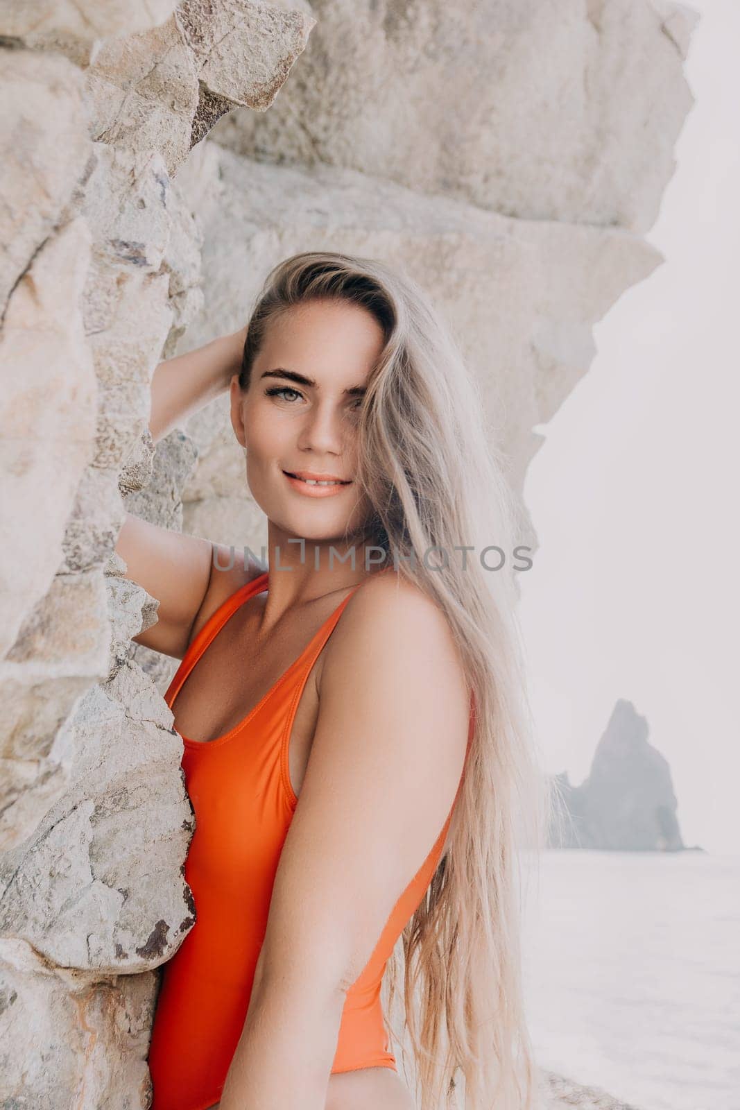 A young woman with long blonde hair is wearing an orange swimsuit and smiling while standing by rocks.